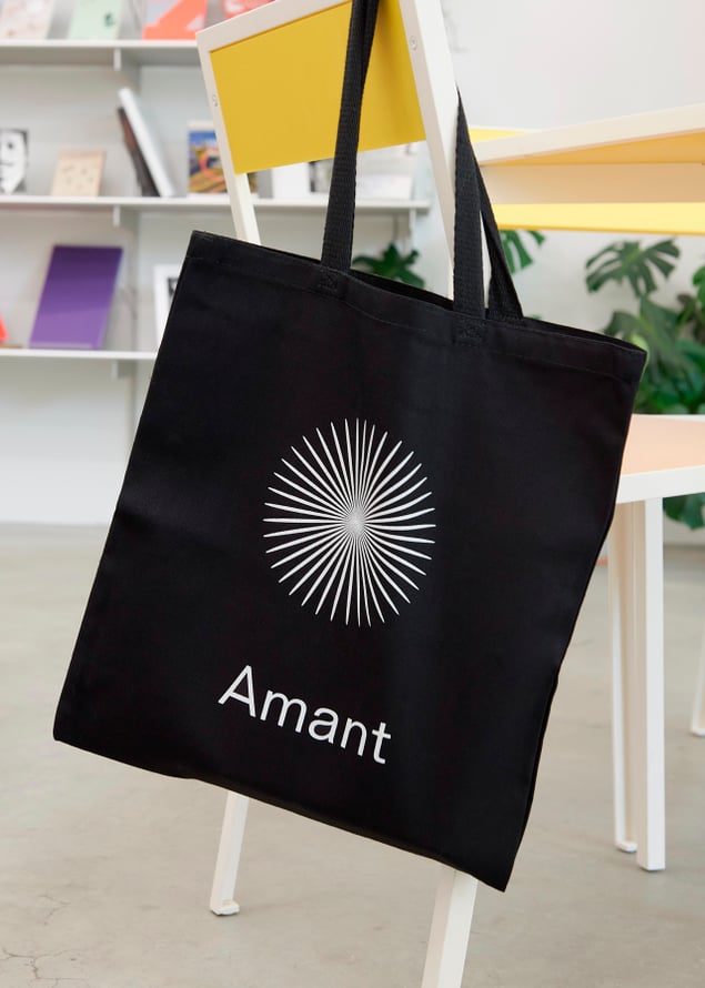 A tote bag with the Amant logo and wordmark hangs off the back of a yellow chair.
